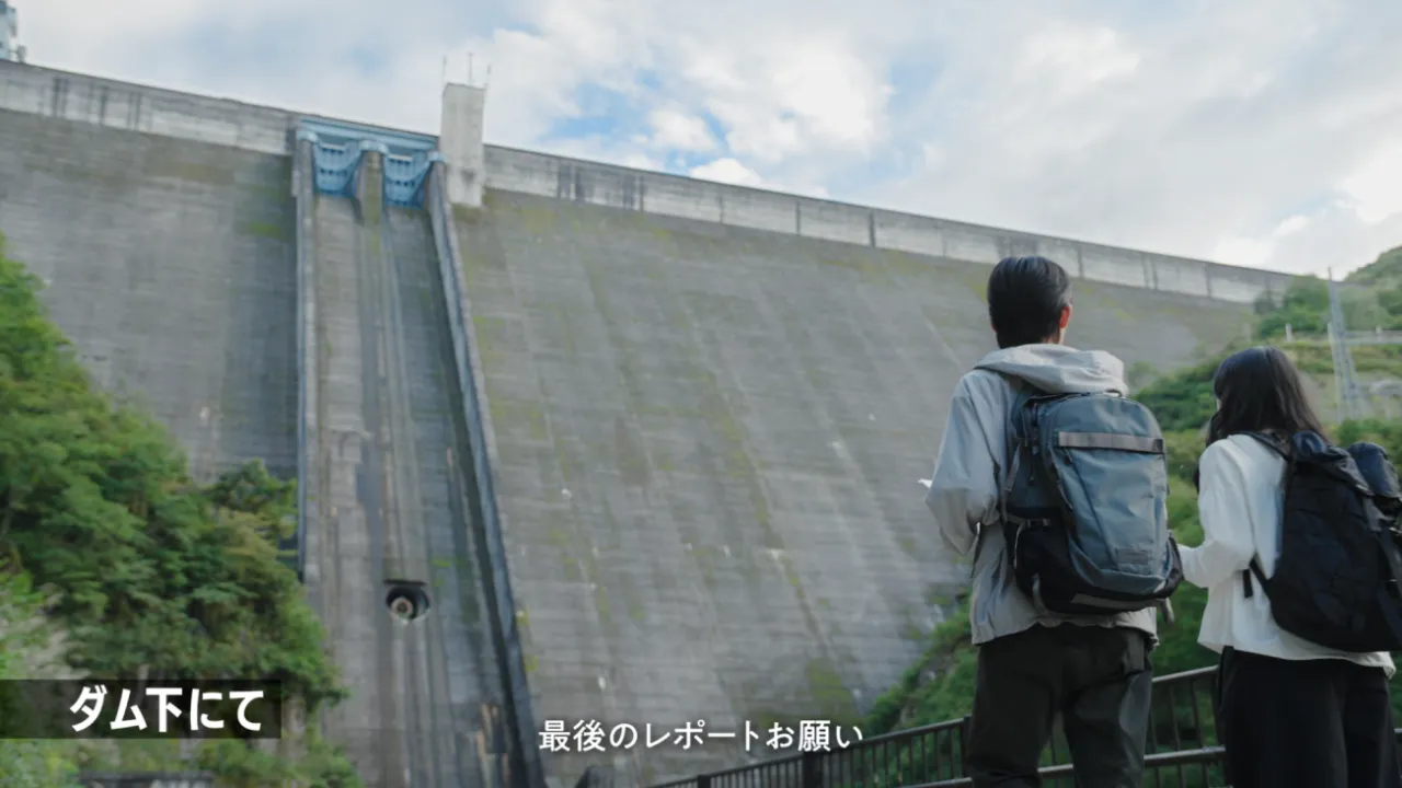 「ダム下にて」の表示と、奥只見ダムを見上げる山時聡真さんと池田エライザさんの後ろ姿