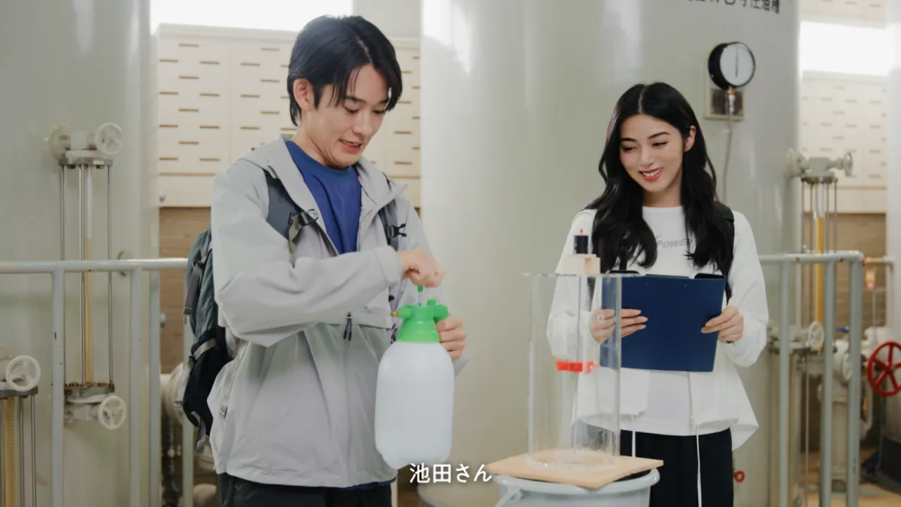 ダムの設備室で器具を手にする山時聡真さんとクリップボードを持つ池田エライザさん